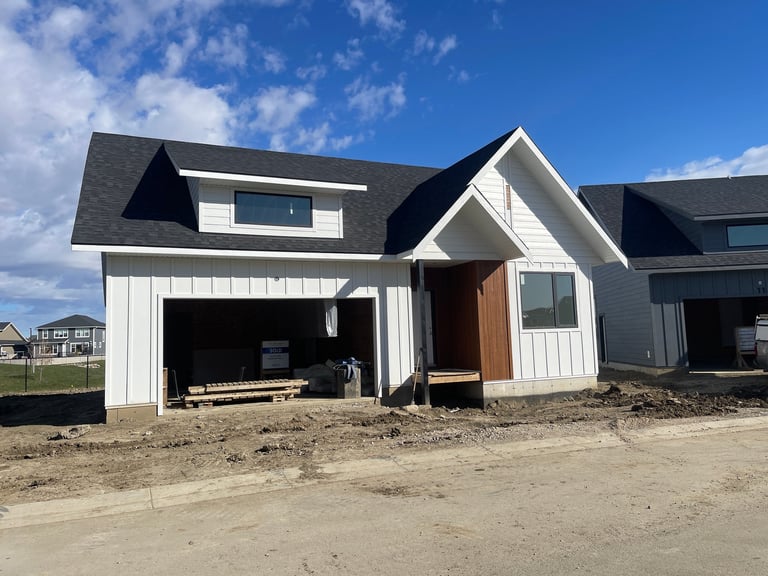 Modern white farmhouse with black roof under construction, featuring open garage door and brown front entrance door, set on sandy lot with blue sky background