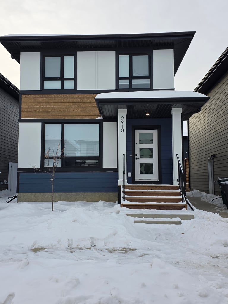 Modern two-story house with black and white exterior, wood accents, and large windows covered in snow during winter